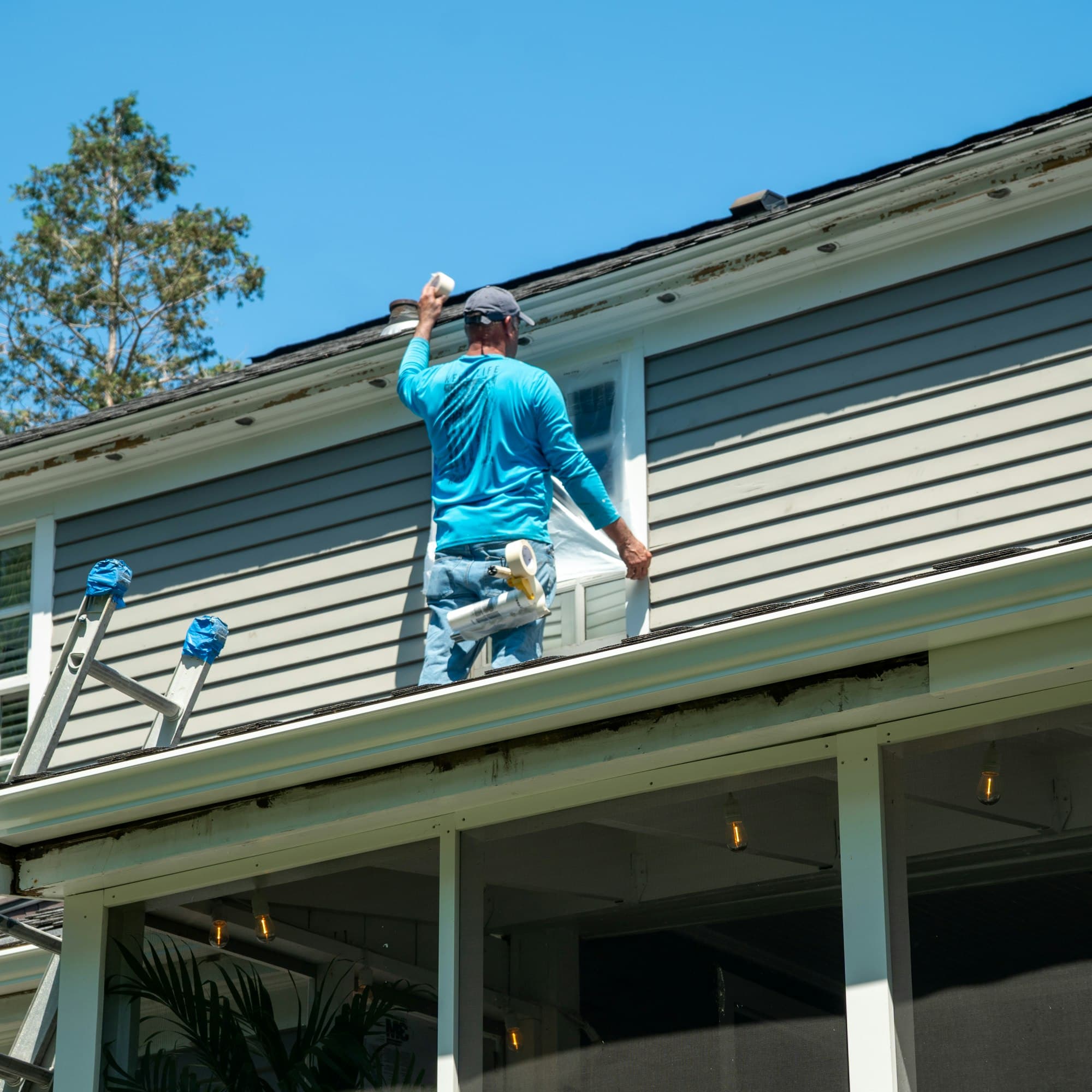 A worker on the roof of a two-story house with gray siding, under a clear blue sky